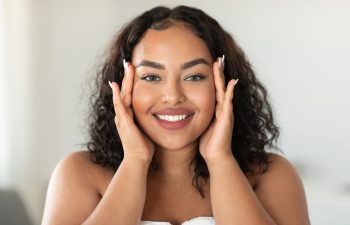 Beautiful black body positive woman touching her face and smiling at camera, making beauty treatments at home. Young african american lady enjoying selfcare